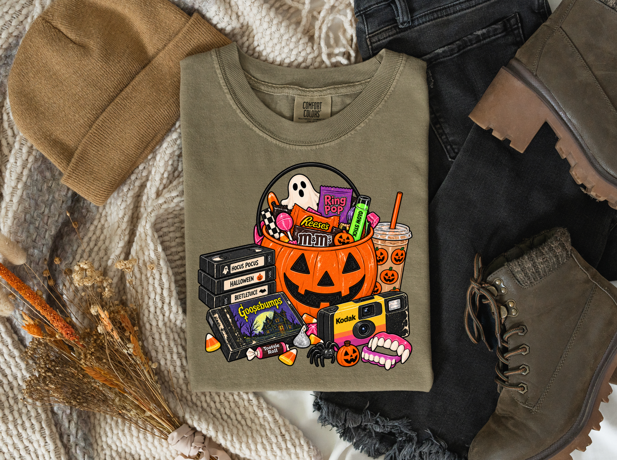 Halloween nostalgia graphic featuring a pumpkin candy bucket, VHS tapes, a camera, and spooky treats on an olive tee